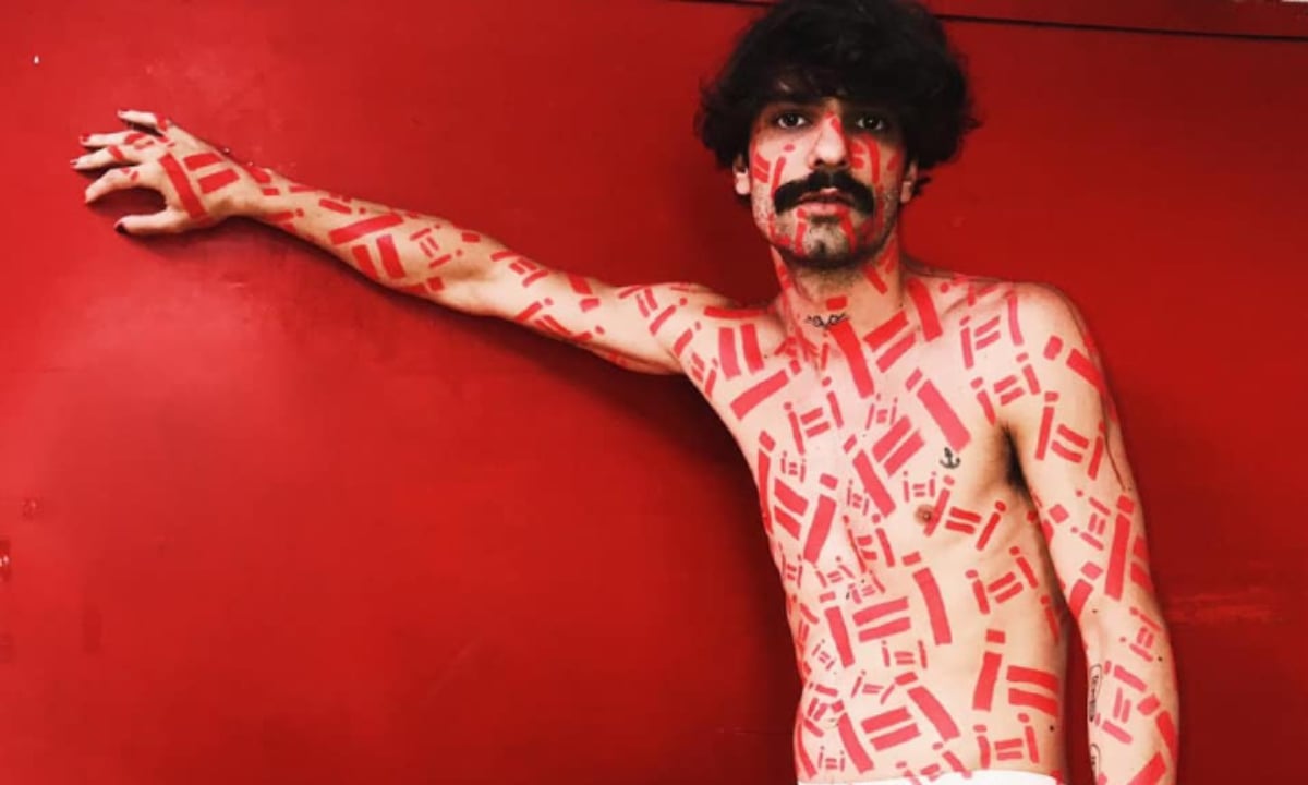 Vinicius Couto com o corpo carimbado com o símbolo I=I diante de um fundo vermelho